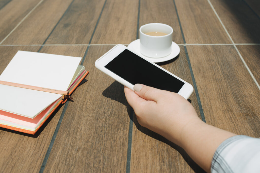 hands holding white mobile phone with blank black desktop screen with notebook and coffee cup on wooden table in cafe