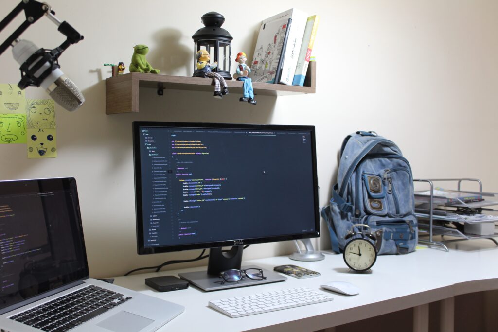 Monitor sitting on desk with alarm clock, laptop backpack, and small office supplies