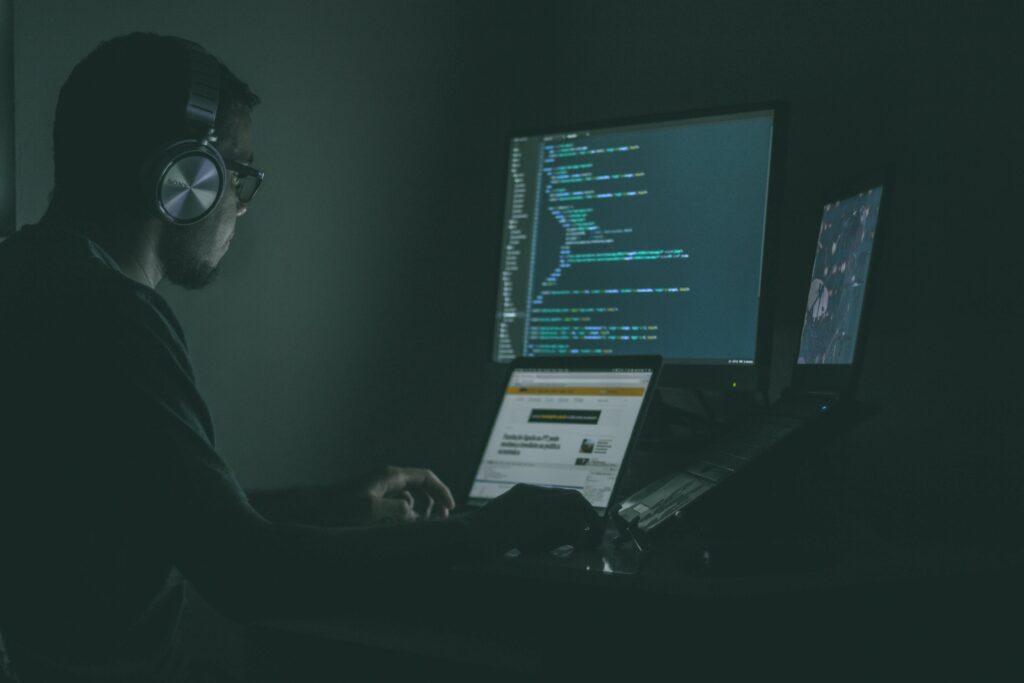 Person sitting in dark room with laptop and two monitors.